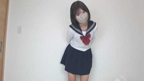 Video of a young Asian woman with short black hair, wearing a Japanese schoolgirl uniform with a red bow, gagged, and tied up against a plain white wall.