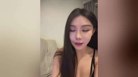 A video of an East Asian woman with long, straight black hair and light skin, wearing a black spaghetti strap top, sitting on a beige couch in a dimly lit room with gray curtains.