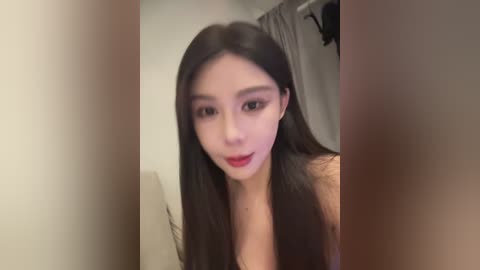 A video of an East Asian woman with long black hair, fair skin, and a slender physique, wearing a sleeveless top, smiling slightly. The background shows a beige wall and part of a gray curtain.