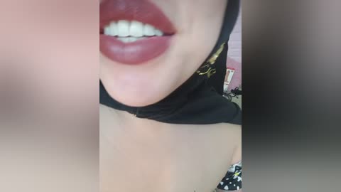 Close-up video of a woman with fair skin, full red lipstick, and a black choker. The background is blurred, featuring a pink object and a patterned fabric.