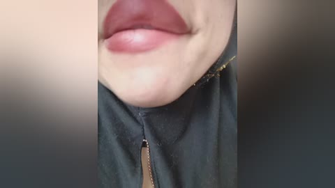 A close-up video of a person's lower face, showing full lips with a glossy, deep red lipstick and slightly parted, revealing a hint of teeth. The person is wearing a dark green jacket with a zipper. The background is blurred, making the lips and jacket the focal point.