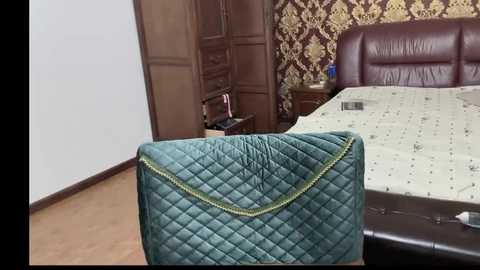 Video of a cozy bedroom with a dark brown headboard, beige quilted bedspread, green quilted cushion, brown wooden doors, and ornate wallpaper.