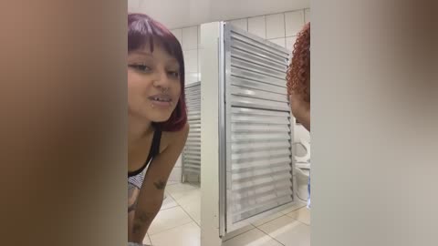 Video of a young woman with medium brown skin and straight, shoulder-length red hair, wearing a black and white striped bra, leaning over in a tiled bathroom with a metal door in the background.