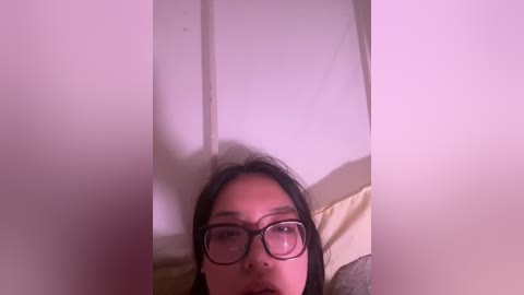 A close-up video of a young woman with long black hair and glasses, lying on her back with a beige pillow. The background is a plain, light-colored wall.