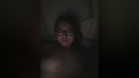 A dimly lit video captures a young Asian woman with long black hair, glasses, and medium-sized breasts, lying in a bed with white pillows, partially obscured by shadows.