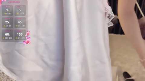 Video of a woman's lower body in a white lace dress with pink floral embroidery, standing beside a white table. A digital display on the left shows vital signs and health metrics.