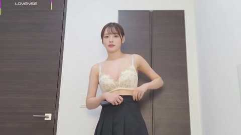 A video of a young Asian woman with straight black hair, wearing a beige lace bra and black skirt, standing in front of a wooden door.