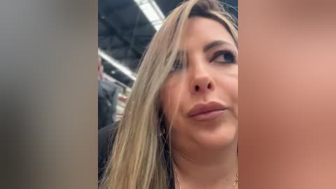 A video shows a close-up of a young woman with long, straight blonde hair, fair skin, and full lips, looking up. The background is blurred, possibly an indoor setting.