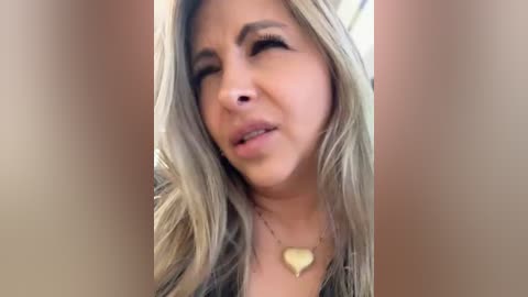 Video of a blonde woman with long hair, wearing a heart-shaped necklace, winking, with a neutral background.