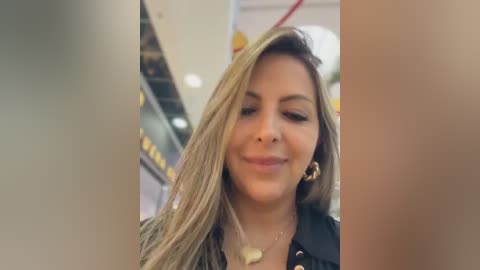 Video of a smiling woman with long blonde hair, wearing a black jacket and gold hoop earrings, indoors, blurry background.