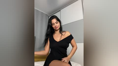 Video of a Latina woman with long black hair, medium skin tone, wearing a black off-shoulder dress, sitting on a bed in a modern, minimalist bedroom with white and grey walls.
