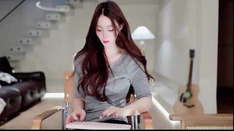 Video of an Asian woman with long, wavy brown hair, wearing a grey top, seated in a wheelchair, reading a book, in a modern, minimalist living room with white walls, a staircase, and a guitar.