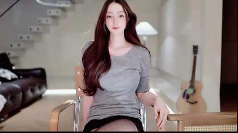 Video of a woman with long, straight brown hair, wearing a grey t-shirt, sitting in a modern, minimalist living room with a staircase, guitar, and white lamp.