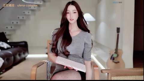 Video of an East Asian woman with long brown hair, wearing a gray dress, sitting on a wooden chair, holding a book, in a modern, minimalist living room with a staircase, guitar, and a lamp.
