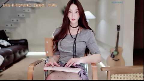 A video of an East Asian woman with long brown hair, wearing a grey sweater and a black choker, seated at a wooden desk in a modern, minimalist living room.