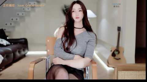 Video of an Asian woman with long dark hair, fair skin, and a slender figure, wearing a grey sweater and black skirt, sitting in a modern living room with a wooden chair and a guitar.