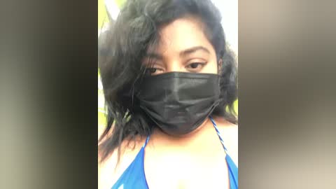 A close-up video of a South Asian woman with medium brown skin, wearing a black face mask and a blue halter top, her long black hair partially covering her face.