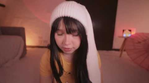 A video of a young woman with long black hair and a white knit beanie, wearing a yellow shirt, sitting indoors. Background includes a grey couch, a pink parasol, and a glowing lamp.