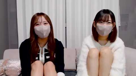 Video of two Asian women, both wearing face masks, sitting on a couch with beige cushions, dressed in casual attire; one in a black sweater, the other in a white fuzzy jacket.