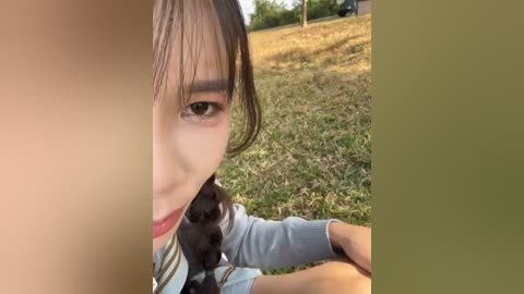 Video of a young Asian girl with long black hair and a light blue sweater, sitting on grass, partially obscured by a large, light-skinned hand. Background features a grassy field and trees.