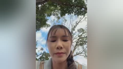 A video of a young Asian woman with long, dark hair, wearing a striped shirt, looking up at a bright blue sky with fluffy white clouds and lush green trees.