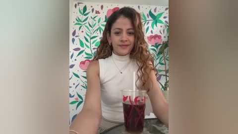Video of a young woman with wavy brown hair, wearing a white sleeveless top, sitting at a table. She holds a red drink in a clear plastic cup. Floral wallpaper in the background.