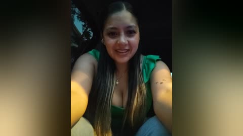 Video of a smiling, light-skinned woman with long, straight black hair and light brown highlights, wearing a green top, taking a selfie outdoors.
