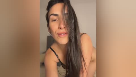 Video of a young woman with long, straight dark hair, light skin, and a confident smile. She's wearing a black bra, leaning forward with her hands on the ground, in a blurred bedroom setting.
