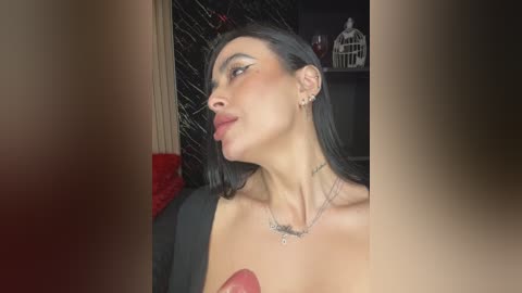 Video of a young woman with long black hair, wearing a black top and a silver necklace, standing in a dimly lit room with dark walls and a birdcage.