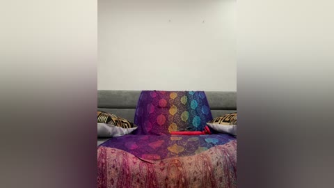 Video of a minimalist, gray couch with a colorful, abstract-patterned throw blanket featuring vibrant hues of purple, pink, and blue. The background is a plain, white wall, and two patterned pillows are placed on either side of the blanket.
