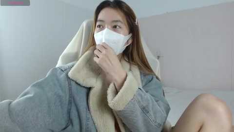 Video of an Asian woman with long brown hair, wearing a white medical mask, light gray sweater, and beige fur-lined jacket, sitting on a white chair in a minimalist, well-lit room.