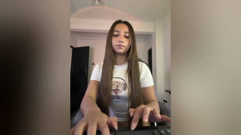 Video of a young Latina woman with long brown hair, wearing a white t-shirt with a graphic design, sitting at a computer desk in a modern, minimalist room with white walls and a black door.