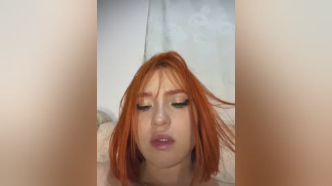 Video of a young woman with short, bright red hair and fair skin, wearing minimal makeup, lying down with her eyes closed and lips slightly parted. Background features a light-colored curtain.