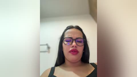 Video of a plus-size woman with long black hair, wearing black-framed glasses and a black tank top, standing in a bathroom with beige walls and a toilet paper dispenser visible.