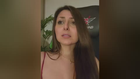 Video of a young woman with long brown hair, wearing a red spaghetti strap top, standing indoors with a green plant and a black gaming chair in the background.