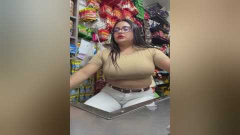 Video of a plus-sized woman with long black hair, wearing glasses and a beige crop top, sitting in a cluttered convenience store aisle, surrounded by colorful snacks and chips.