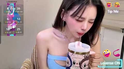 A young Asian woman with fair skin and straight black hair, wearing a light blue strapless top, drinks from a pink and white cartoon-themed cup. Background shows a TV screen displaying Livense.com's streaming interface.