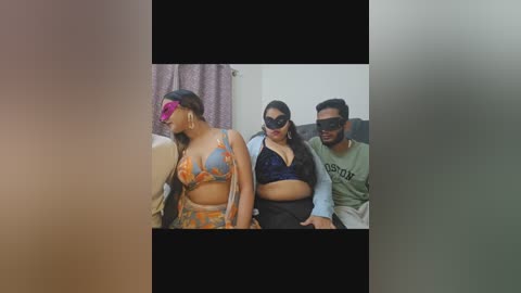 Video of three individuals in a dimly-lit room, dressed in colorful, patterned costumes. The woman in the center wears a blue bra and has a dark mask. The man on the right wears a green shirt with a black mask.