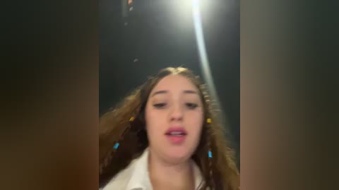 A blurred video of a young woman with long, wavy brown hair, wearing a white top, standing against a dimly lit background.