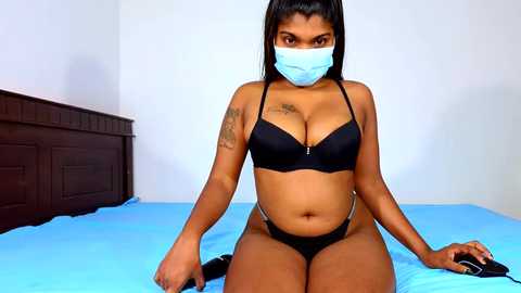 Video of a young Black woman with medium brown skin, wearing a black bra and panties, sitting on a blue bed, wearing a mask, in a minimalistic bedroom setting.
