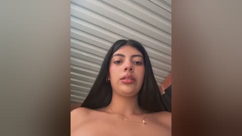 A video of a young woman with long, straight black hair, light brown skin, and full lips. She is topless, wearing a delicate gold necklace, standing in a room with a white ribbed ceiling.