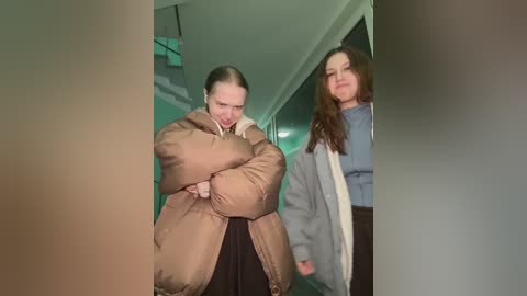 Video of two women in a dimly lit hallway. One wears a brown puffer jacket, black skirt, and holds a white bag. The other, in a grey sweater, has long brown hair and a white bag.