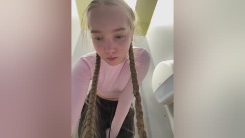 Video of a young Asian woman with light skin and long, braided hair, wearing a pink long-sleeve top and plaid skirt, standing in a public restroom stall.