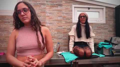 Video of two women in a modern, tiled bathroom. One sits on a counter, wearing a pink crop top and glasses, the other, a brunette in a beige sweater and black leggings, sits on a stool.