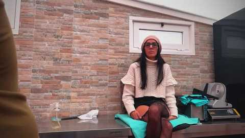 Video of a woman with long black hair, wearing glasses and a pink beanie, seated on a teal mat in a modern, beige-stone tiled room with a window and black furniture.
