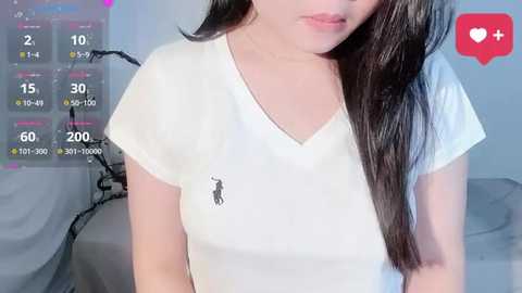 Video of an Asian woman with long, straight black hair, wearing a white Polo Ralph Lauren T-shirt, in a minimalist bedroom with grey walls and a bed.