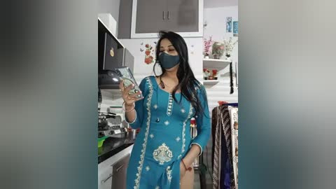 A video of a South Asian woman in a teal sari, wearing a face mask, taking a selfie in a modern kitchen.