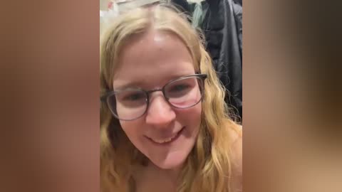 Video of a smiling, fair-skinned woman with wavy, shoulder-length blonde hair, wearing glasses, and a light-colored top. Background shows blurry, dark clothing. The image is slightly out of focus.