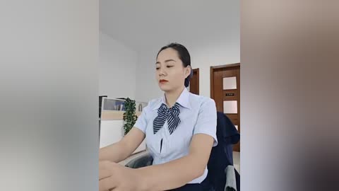 Video of an Asian woman in a light blue button-up shirt with a striped bow tie, seated at a desk in a minimalist office.