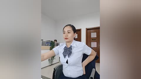 Video of a young East Asian woman with long black hair tied back, wearing a white button-up shirt and blue and white striped tie, sitting in a chair in a modern office with a wooden door and green plants in the background.